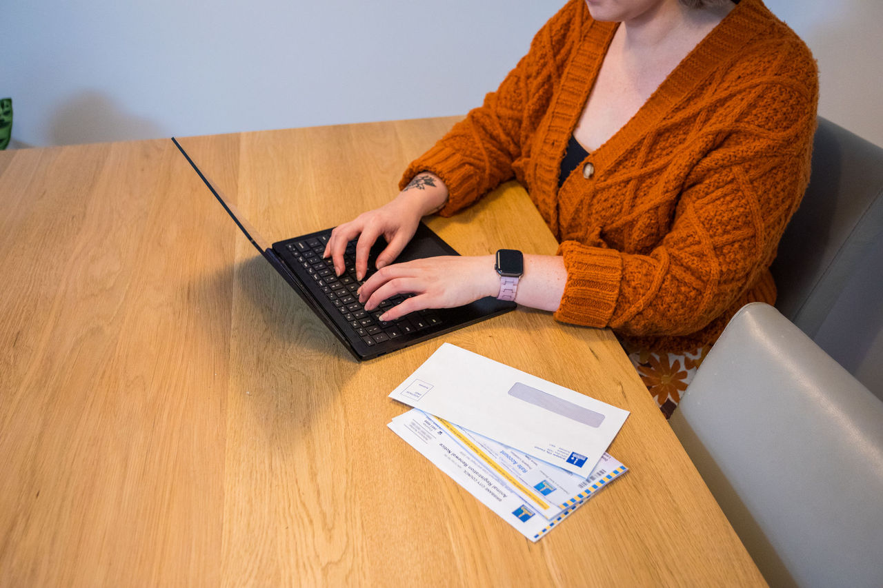 Close-up of a woman from neck down with a laptop on a kitchen table and a rates notice beside the computer.