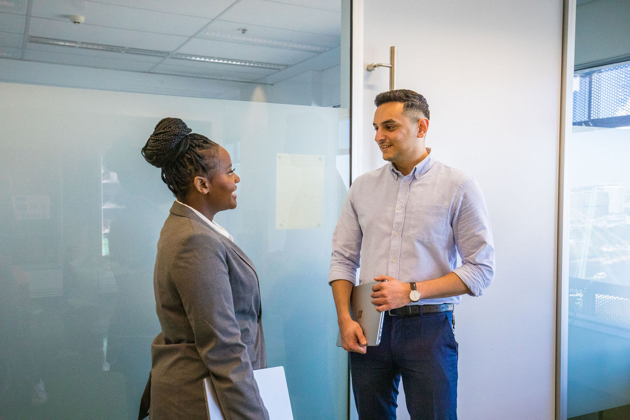 Female and male office walkers standing in a work foyer talking. They both have laptops under their arms.