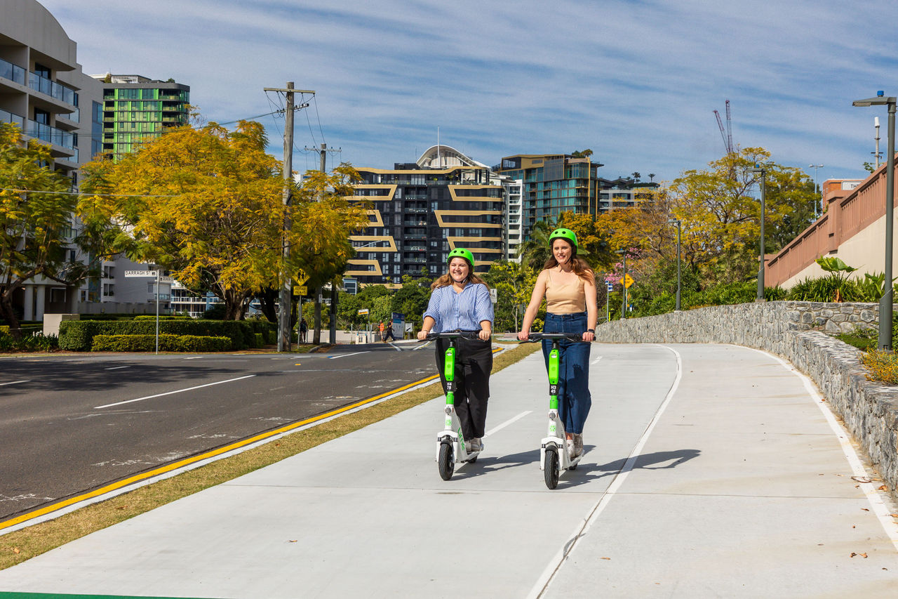 Two women on e-scooters on a bikeway at Kangaroo Point with apartment buildings in the background.