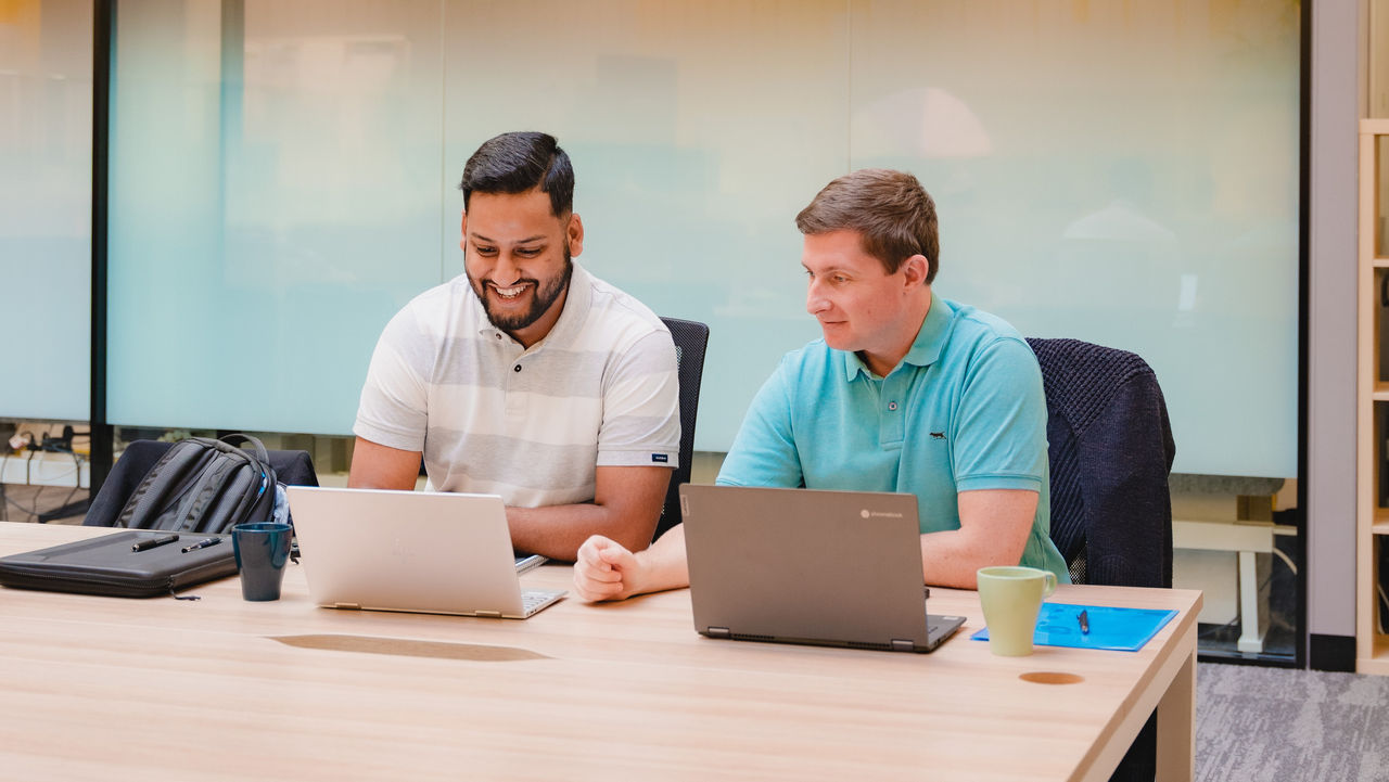 Two businessmen in casual business attire working together on laptops in an open workspace.
