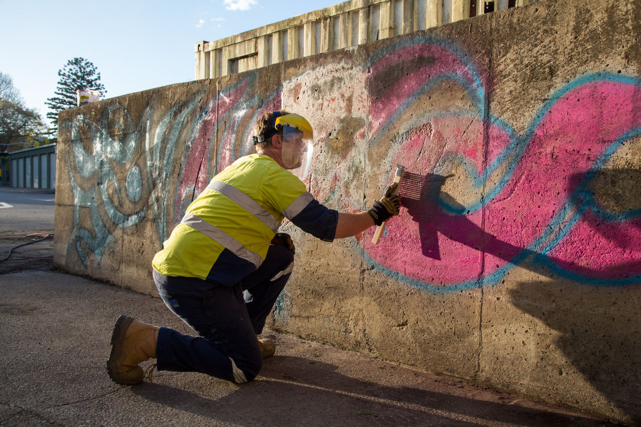 Man wearing yellow vest and protective mask cleaning graffiti from a wall.