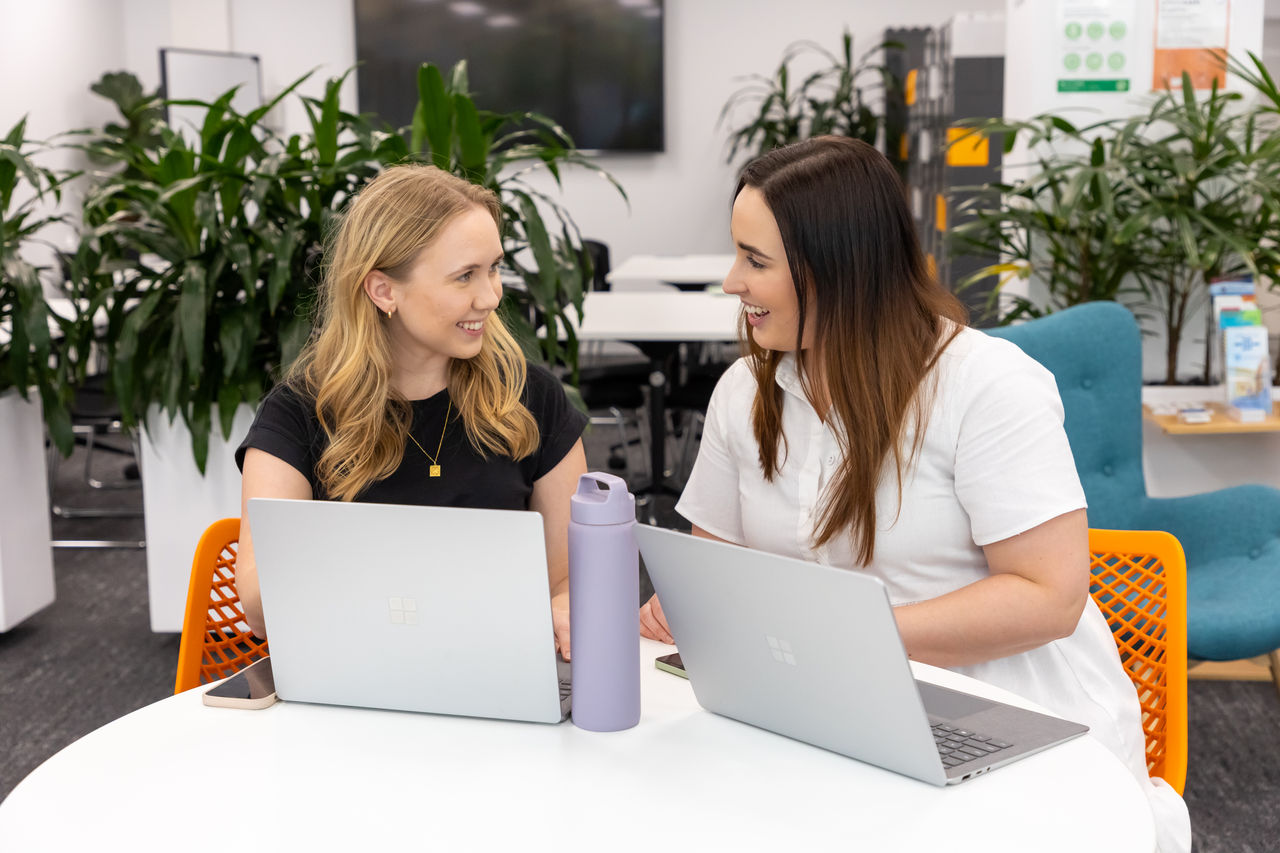 Two young women working with laptops in front of them on a round business table in an open office setting.