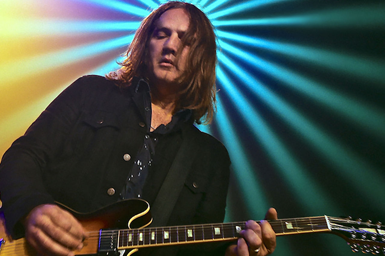 Ian Haug - mentor for The QUBE Effect 2026. Shot inside with bright coloured lighting behind subject. Ian is playing a guitar.