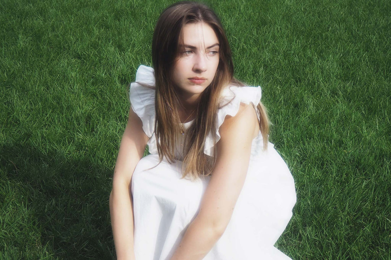 Maddison Kate in a white dress, crouching down, on a lawn. Finalist in The QUBE Effect 2026.