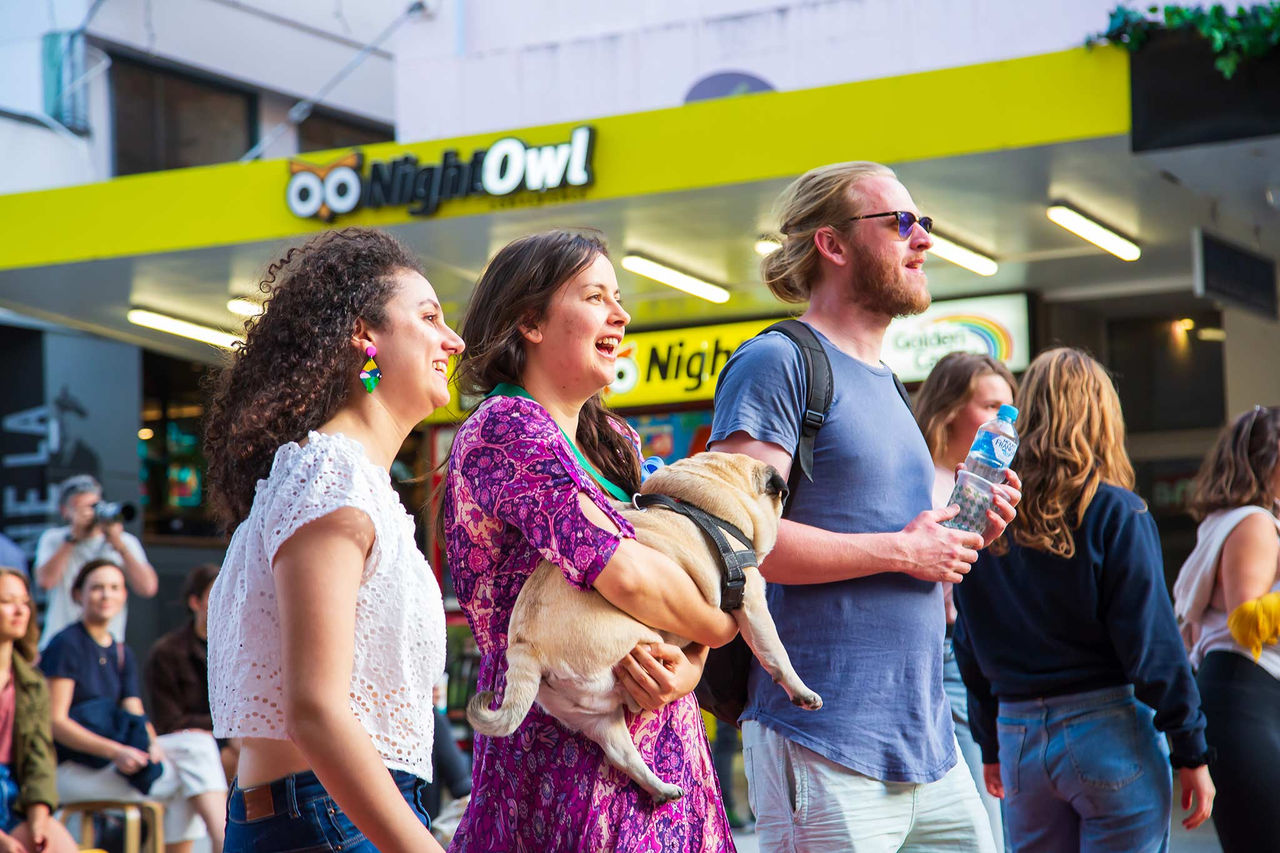 Three people watching an event in Brunswick Street Mall. Middle lady is cradling a pug dog in her arms.