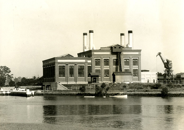 This black and white photograph of the New Farm Powerhouse was taken from the river circa 1930. The large brick and concrete industrial building looks like 2 large cubes with one slightly higher than the other. It has large regularly spaced windows, and 4 large steel chimneys on the roof. The building sits on land near the riverfront. There is a crane in the background.