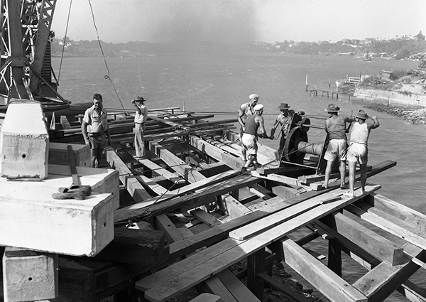 This black and white photograph shows the Norman Creek Bridge construction in the 1950s. Taken from the elevated construction site, it shows workmen in shorts, singlets and hats on large timber beams and concrete blocks winding a large spool of steel cable. The Brisbane River can be seen below and in the background.