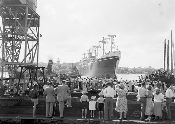 This black and white photograph from 1950 shows the 6 ton freighter 'Binburra' at the entrance to a slipway on the Brisbane River. In the foreground, a large crowd of men, women and children stand on the dockside to watch the ship arrive. Crane scaffolding to the left of the crowd obscures what could be a steam tug boat on the river beside the freighter.