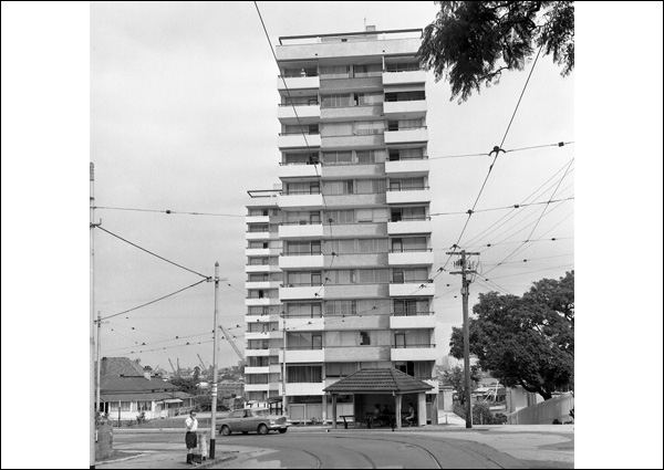 This black and white photograph taken from the middle of a road in 1967 shows 'Gleneagles' apartments comprised of 2 plainly designed high-rise buildings. The buildings have solid masonry balconies with large areas of glazing underneath. In this image, one building is partially visible behind the other. There is a bus shelter in front of the building, tram lines on the road in the foreground and a web of power lines overhead.
