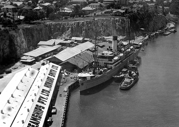 This is a black and white image showing an aerial view of Howard Smith Wharves in 1938. The wharves have large, long, rectangular single storey sheds below a sheer rock cliff-face. There is a large ship, a barge and a tug boat docked the jetty alongside the sheds. One of the sheds has 'Steamships Pty Ltd Howard Smith Ltd' painted on its roof.