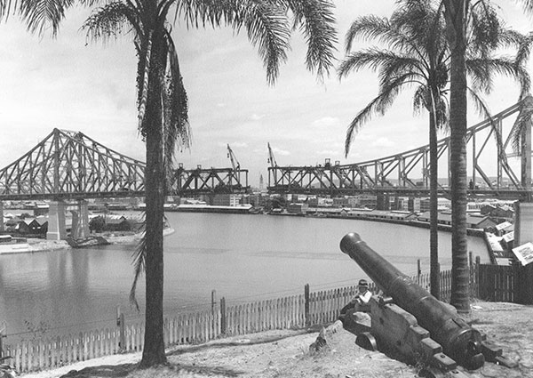 This black and white image shows the Story Bridge construction almost completed in October 1939. The image was taken from a clifftop park and shows both sides of the bridge with an incomplete section in the middle. There are palm trees in the foreground on either side of a cannon that points toward the Brisbane River.