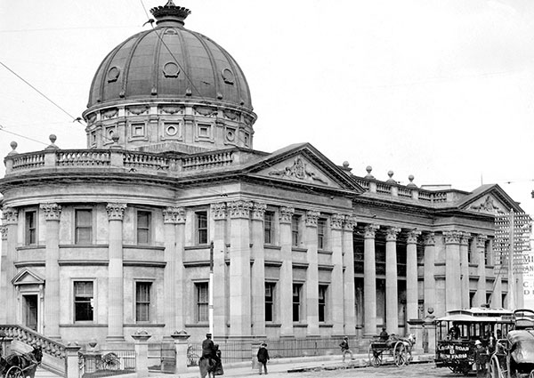 This is a black and white photograph of Brisbane Customs House circa 1890. It shows the 2-storey masonry building with Corinthian columns and pilasters around its circumference and a large double storey dome on the roof. There is a tram or trolley bus and people on horses or in horse-drawn buggies on the street in the foreground.