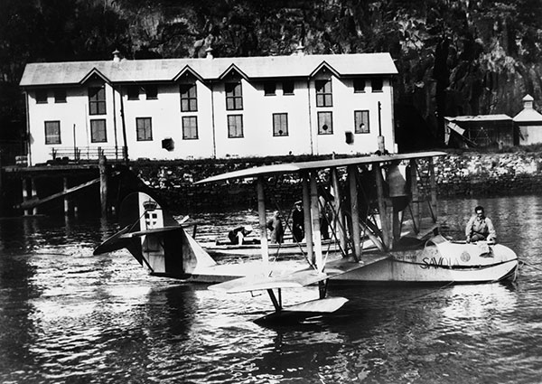 This black and white photograph shows a seaplane floating on the Brisbane River in front of the Naval Stores in 1925. The seaplane has 2 sets of wings stacked one above the other like a bi-plane. It is buoyed on the water by attached floats on the underside of the lower wing tip. A man can be seen standing from the cockpit. The Naval Stores in the background on the riverfront have a white facade with tall windows and decorative, pointed gables.