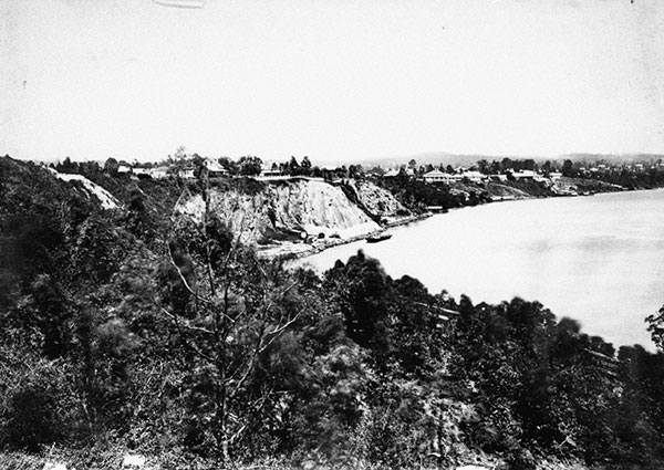This black and white photograph taken from a high location shows the Kangaroo Point Cliffs left of the Brisbane River circa 1890. The light-coloured cliffs are surrounded by dark trees.