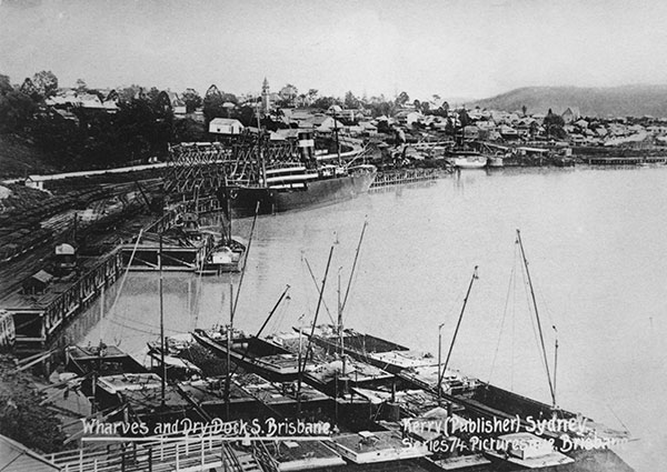 This is a black and white photograph of the wharves and dry dock near the river edge at South Brisbane. It shows several small vessels docked tightly next to each other, and a large cargo vessel in the background.