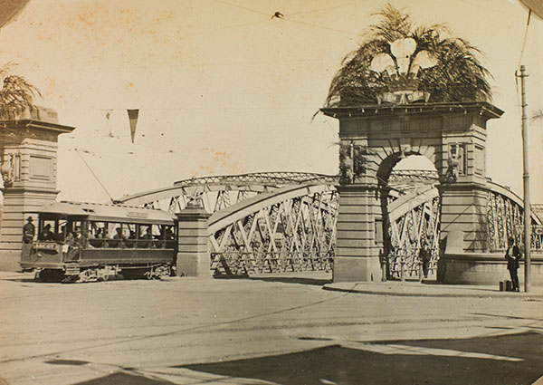 This sepia photograph shows the Victoria Bridge abutment decorated for the visit of Edward, Prince of Wales, Brisbane, 1920. Designed to resemble a traditional stone arch with a central keystone, there are 2 large ornamented archways atop thick pillars at either side of the bridge entrance. These are decorated with palm fronds, fanned upwards and outwards in a semicircle.