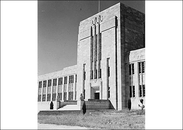This is a black and white photograph of the Forgan Smith Building, University of Queensland, St Lucia, 1950. It shows a tall, imposing building with rectangular detailing on the front and several steps leading to the entrance. The building has lower, 2-storey extensions from either side. The buildings have many evenly spaced windows across the length of each level.