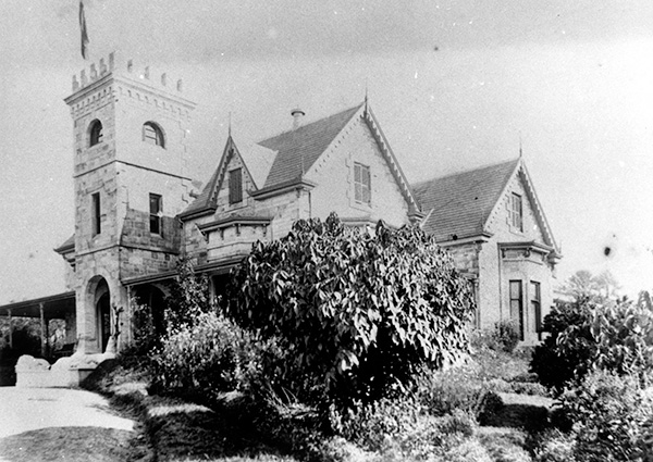 This black and white photograph shows the Hamilton Villa 'Toorak House' after a second storey was added to the house and to entry tower. Informal gardens and shrubs surround the house.