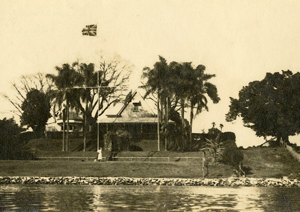 This is an image taken from the river in 1915, and shows Newstead House set up on a slight rise to the riverbank. In front of the house, there are establish palm trees, a Union Jack flag raised full-mast from a flagpole, and several small terraces to the river.