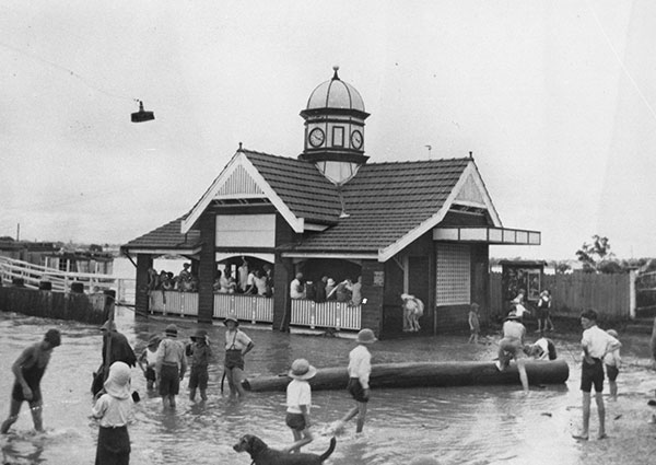 This black and white photographs of the Bulimba ferry terminal shows a quaint timber framed building with a 3-bay verandah under a tiled gabled roof. A low octagonal clock tower with a domed roof site prominently in the centre of the roof. Many people can be seen in the terminal, which is surrounded by floodwaters. Many children and a dog are playing in the water.