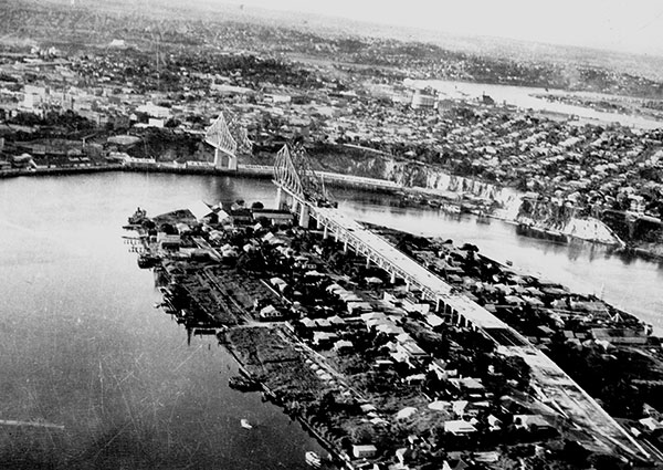 This is a black and white photograph of the Story Bridge under construction. It shows the steel bridge being build from each side to meet in the middle. Situated on the riverbend, each bridge end over the banks is constructed, but its middle expanse is yet to be completed.