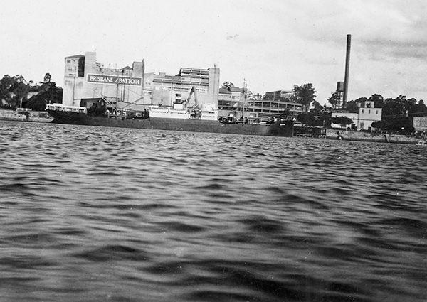 A black and white photograph of the Brisbane River at Colmslie. The water is rippled. On the opposite bank a large barge is docked in front of the Brisbane Abattoir, which is a 4- to 5-storey concrete building.